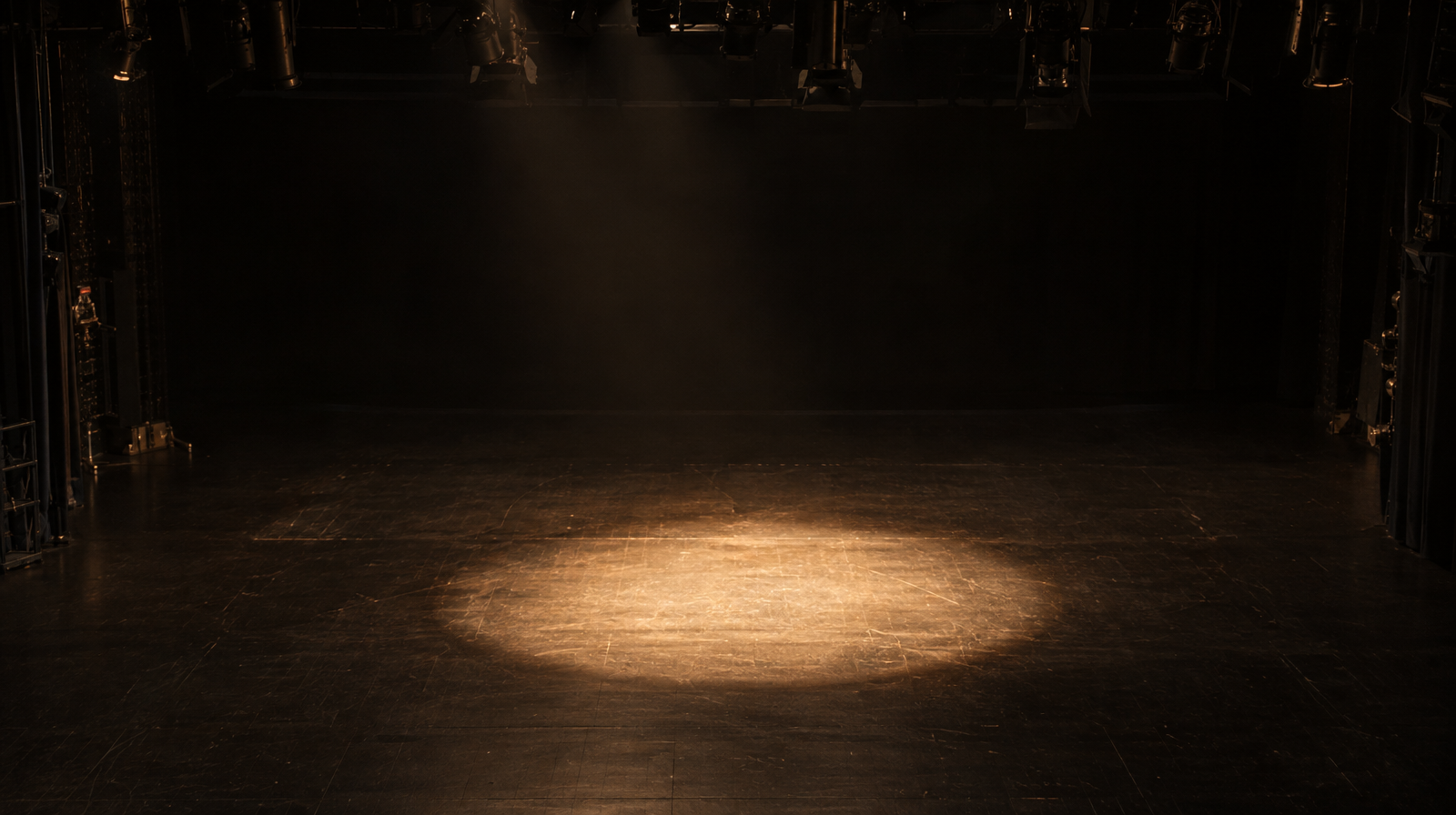 A single motionless spotlight illuminating an empty patch of stage floor, surrounded by dark, retracted stage rigging